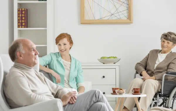 Foto de varias personas mayores sentadas en un salón con una asistente social