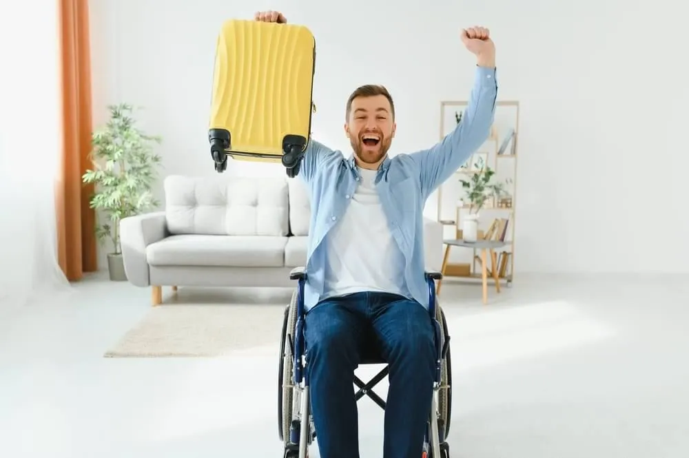 Hombre en silla de ruedas sujetando una maleta en alto, muy alegre, a puto de emprender un viaje