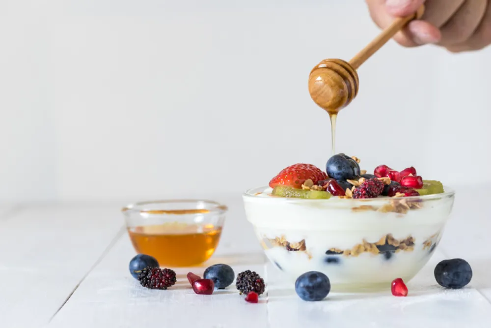 Foto de un mano que vierte miel con un palito en un postre de yougurt y frutas