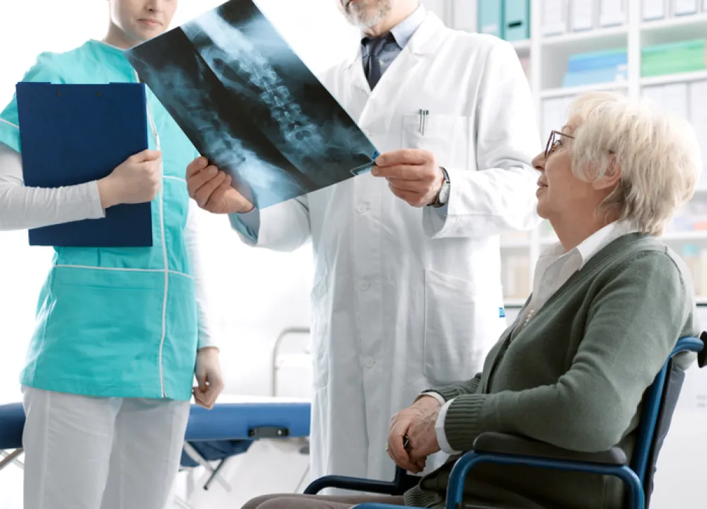 Foto de una persona mayor acompañada de un medico que revisa una radiografía que muestra los huesos de la columna vertebral