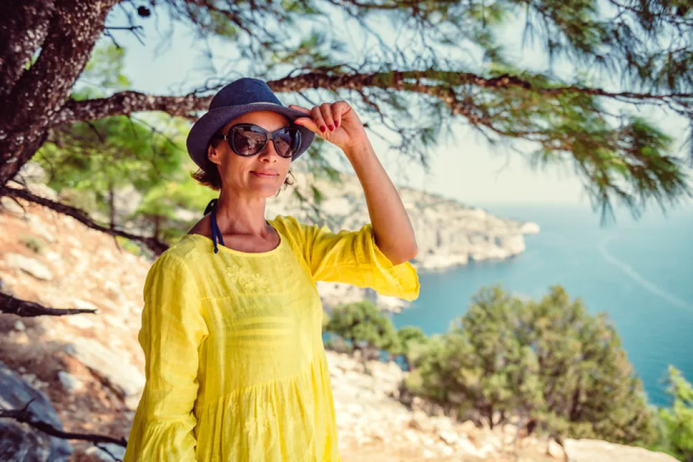 Foto de una mujer adulta con gafas de sol y sombrero debajo de un arbol cerca de una playa