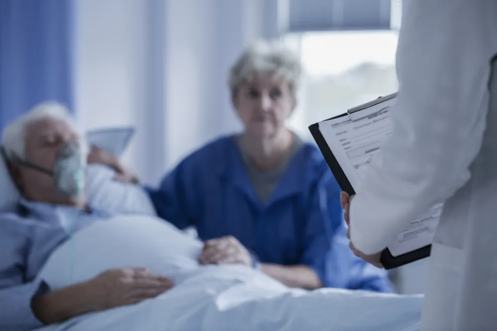 Foto de un hombre tumbado en la cama con oxígeno acompañado de su mujer que escucha a un médico