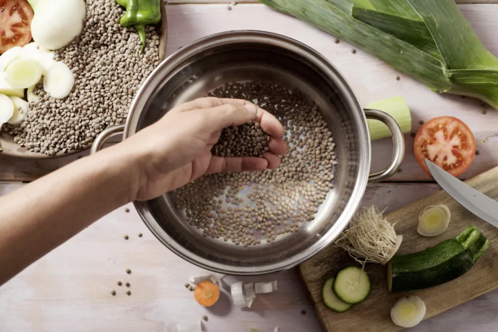 Foto de una mano que prepara en la cazuel un guiso con legumbres y verduras