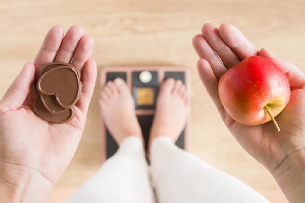 Foto de una mujer subida a una báscula y que sostiene en una mano chocolate y en otra una manzana